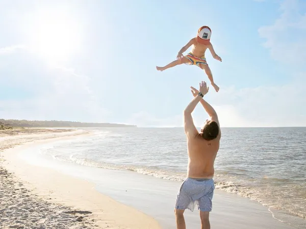 Vater wirft kleinen Jungen am Strand des Campingplatzes Roan Sole Family Camping Village in die Luft.